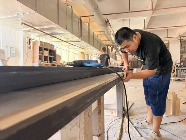 Horace Home worker manually applying edge banding to an approximately 4 m long dark wood veneer panel in the workshop.